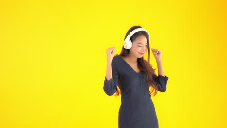 a lovely asian girl wearing bluetooth on-ear headphones is listening to music and dancing on yellow studio background