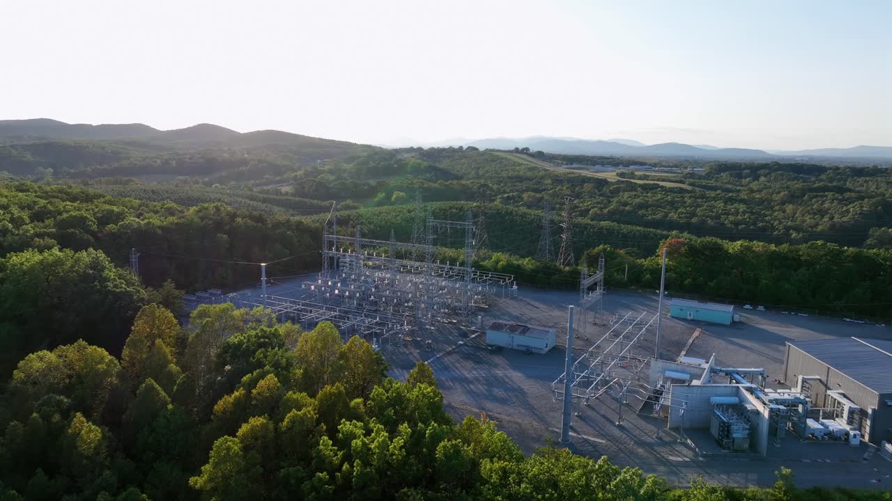Electrical substation with high voltage transmission lines under evening sun in rural American landscape. Drone approaching shot. Green forest landscape in late summer