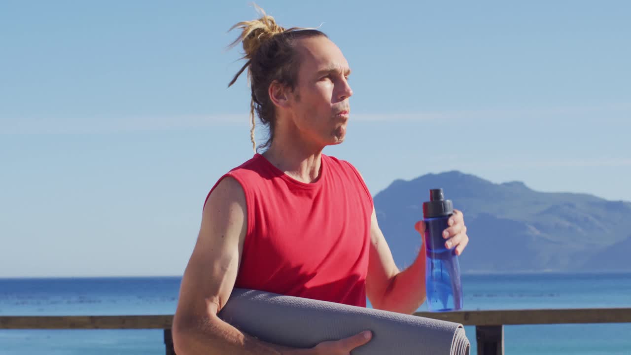 video de un hombre caucásico sonriente con rastas sosteniendo una alfombra de yoga, bebiendo agua al sol junto al mar