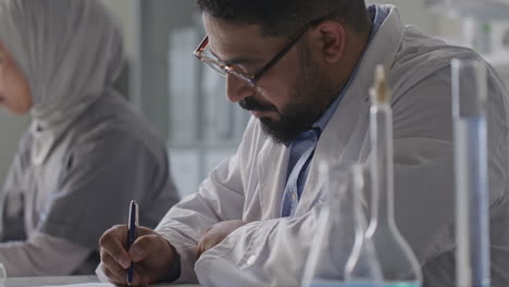 scientist taking notes at laboratory desk next to colleague in hijab