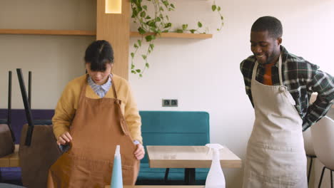 camarera y mesero atando su delantal y luego limpiando la mesa de la cafetería con spray desinfectante y trapo
