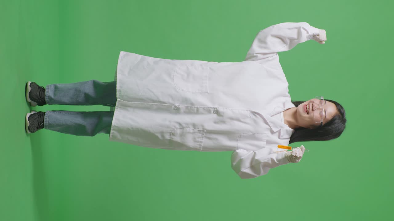 실험관에 오렌지색 액체가 들어있는 아시아 여성 과학자의 몸이 가득 차 있습니다. 실험실의 녹색 스크린 배경에 서있는 동안 미소 짓고 을 구부리고 있습니다.