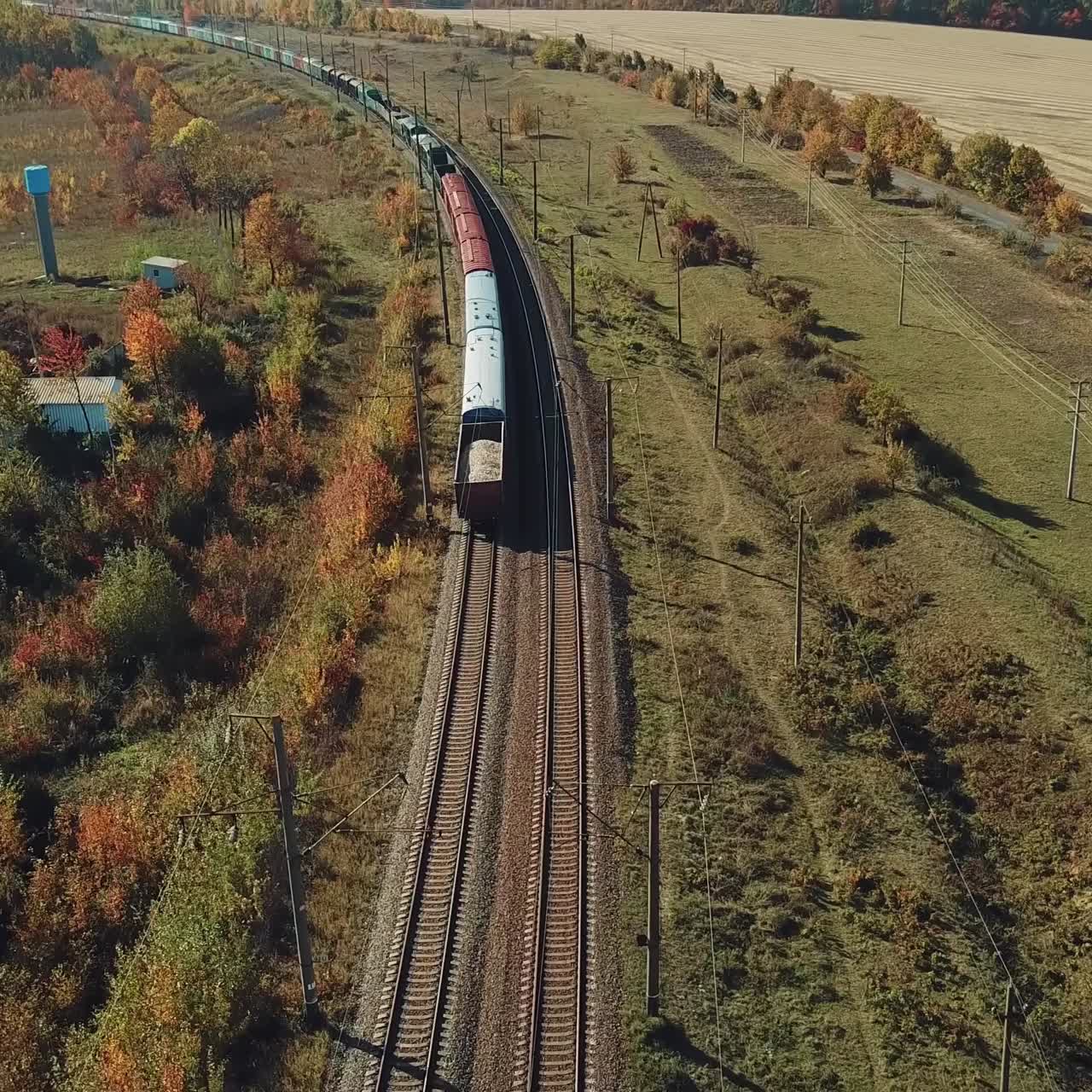 a long freight train with building materials is moving by railway on the background of fields and forests in the summer on a sunny day. Aerial view