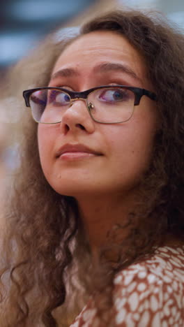 mujer con gafas y cabello rizado mirando a su alrededor en un centro comercial brillantemente iluminado, rodeado de suaves luces bokeh, con detalles de fondo borrosos de las luces del centro comercial y la gente