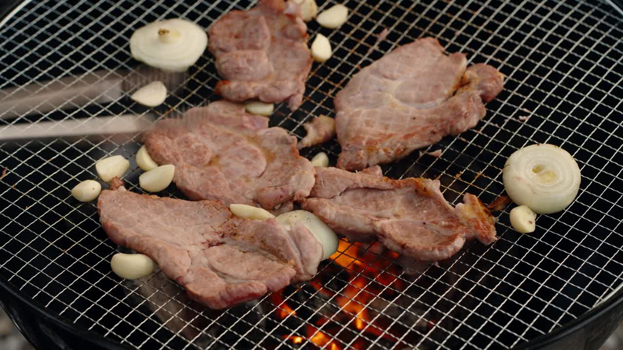 A close-up of thin slices of pork, whole cloves of garlic, and onion rings sizzling on a wire mesh over hot charcoal, a classic style of outdoor Korean barbecue (gogi-gui)