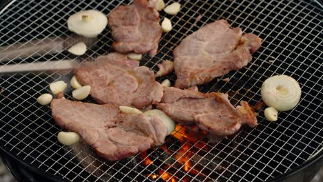 a close-up of thin slices of pork, whole cloves of garlic, and onion rings sizzling on a wire mesh over hot charcoal, a classic style of outdoor korean barbecue (gogi-gui)