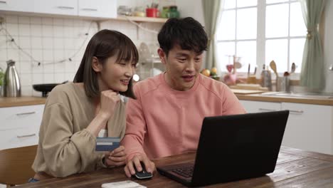 smiling asian husband and wife pointing at computer while discussing what to buy on the internet at home. they pay online with a credit card