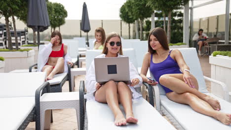 gorgeous girls sunbathe on the deck chairs. ladies at foreground look at laptop. friends behind talk and laugh. spa zone relax.