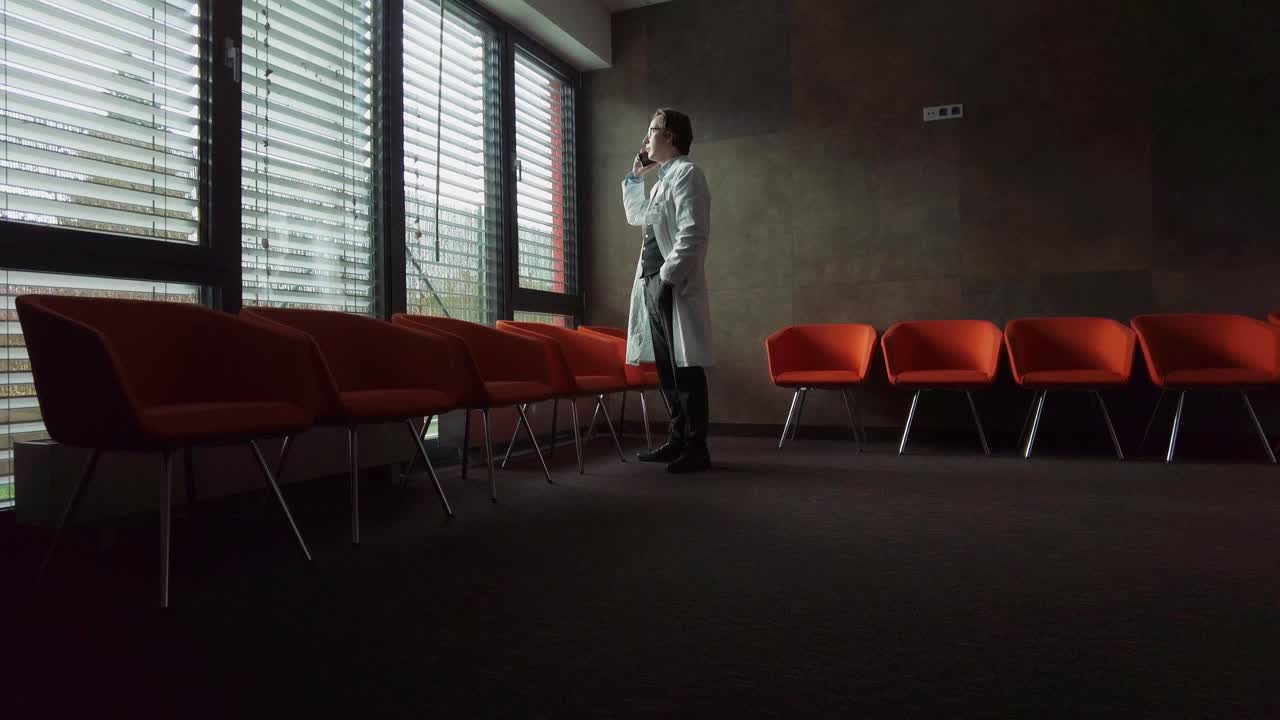 ein junger therapeut im weißen kittel telefoniert mit einem patienten über einen neuen termin in einem raum mit bunten sesseln und licht, das durch die jalousien scheint