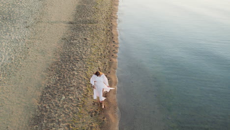 Aerial-tracking-of-young-woman-walking-down-a-sandy-tropical-ocean-beach-at-sunset-or-sunrise,-turns-around---high-angle-slow-motion