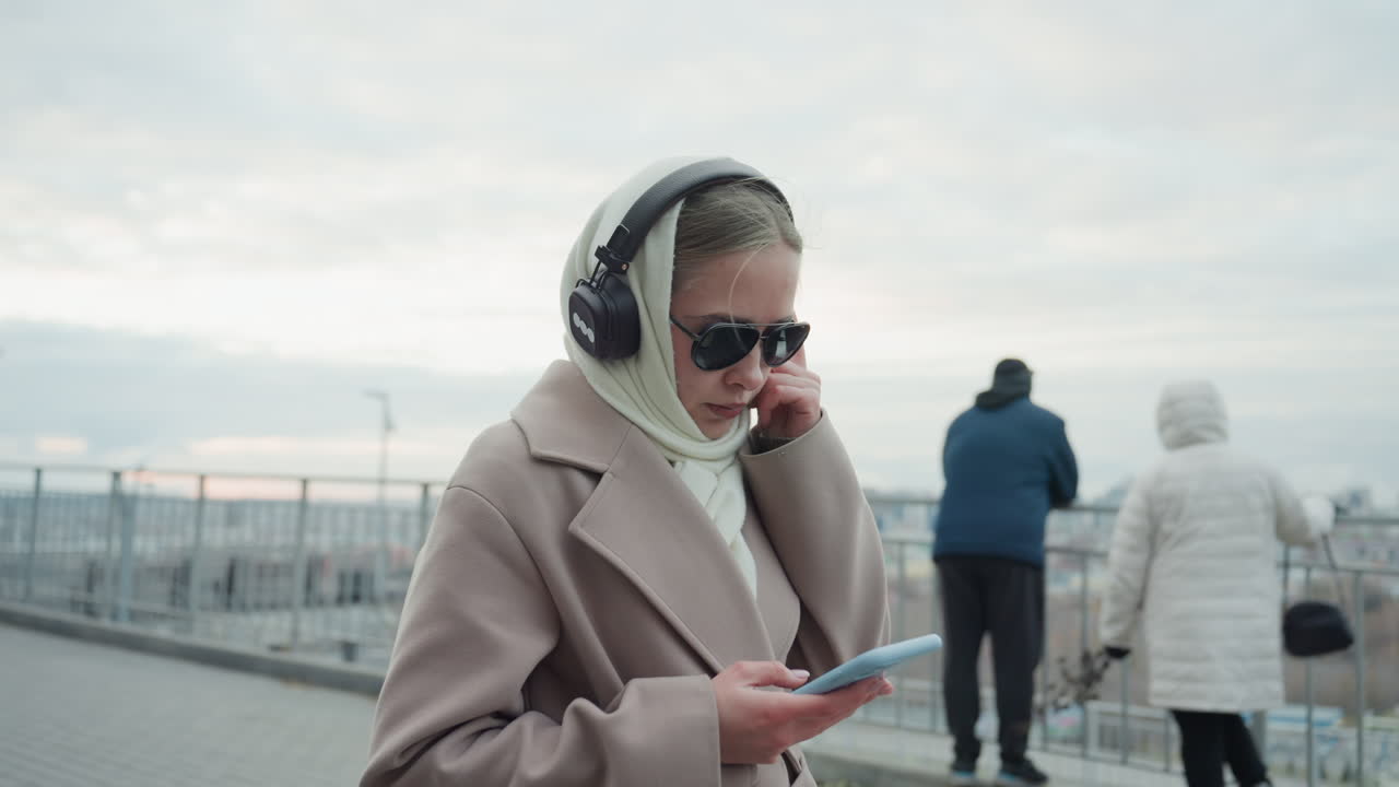 Una giovane donna controlla i messaggi sul telefono su una passeggiata lungofiume, con cuffie e cappotto. Scorre e si ferma per osservare lo skyline, mentre i pedoni in lontananza e il cielo coperto e attenuato creano un'atmosfera urbana tranquilla.