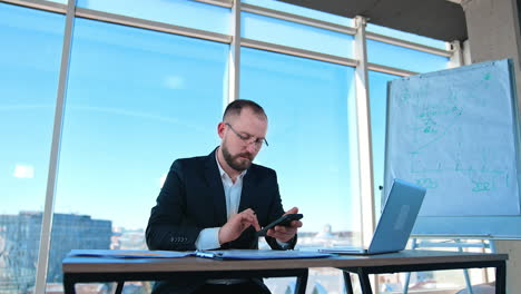focused businessman uses smartphone in workplace. bearded entrepreneur in suit and glasses sitting at table and working next to large window.
