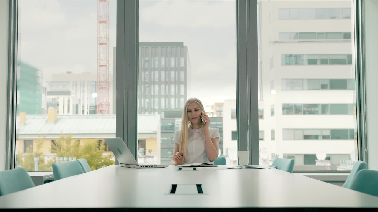 Businesswoman waving with hand while speaking on phone. Stylish businesswoman having phone call while sitting at table in conference hall and waving with hand greeting colleague