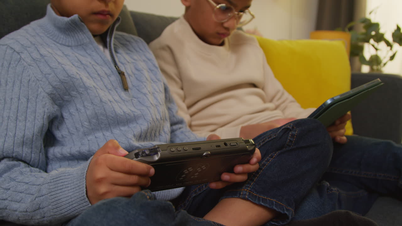 dos niños pequeños sentados en el sofá en casa jugando juegos o transmitiendo a una tableta digital y un dispositivo de juego portátil 4