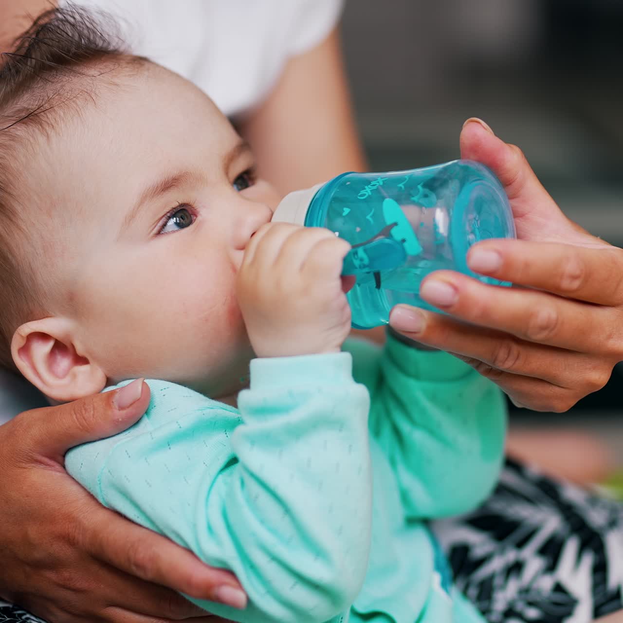 Thirsty little beautiful kid drinking water from a bottle. Caring mom supporting her child and bottle for him. Close up