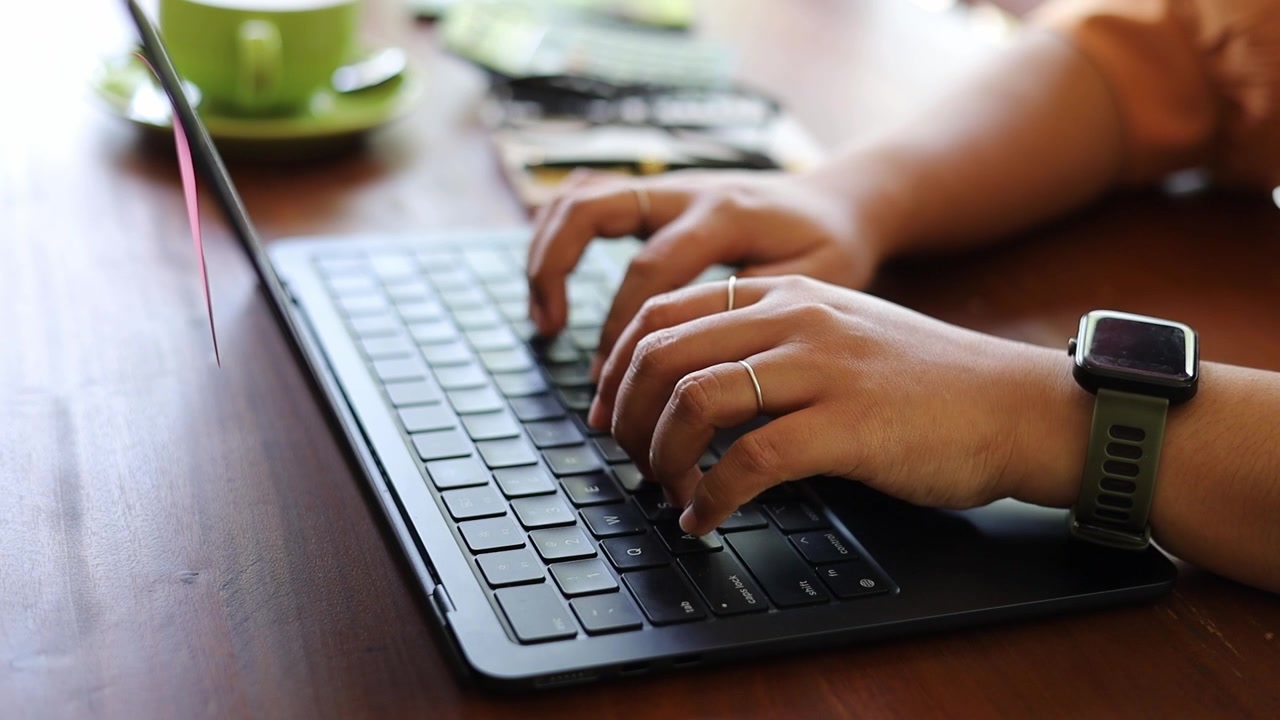 Primo piano di mani femminili d'affari che digitano sulla tastiera di un laptop, scrivono appunti, lavorando in un caffè.