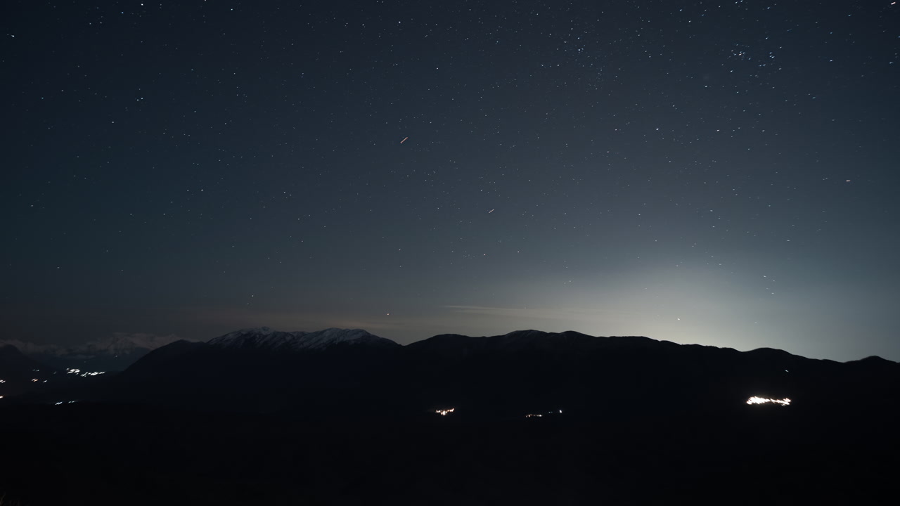 A serene night sky filled with stars over silhouetted mountains, with faint city lights glowing in the distance. Perfect for use in nature documentaries and travel content.