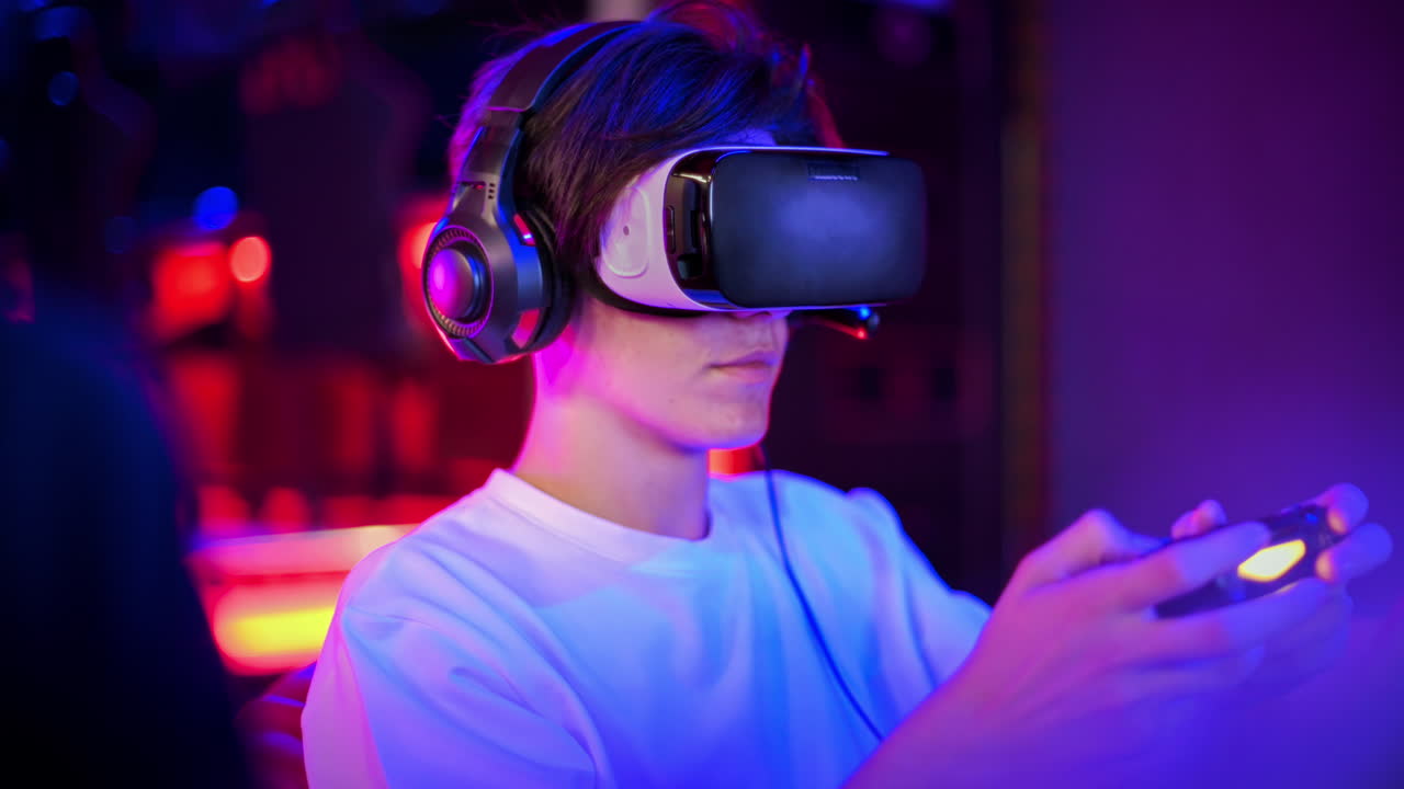 Due amici adolescenti parlano e giocano con una console di gioco in VR con visore e cuffie, usando gamepad mentre sono seduti su pouf. Illuminazione blu e rossa