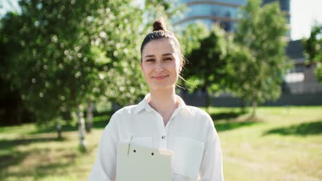 joven mujer de negocios confiada con camisa blanca sosteniendo un clipboard de pie en el jardín verde por el edificio de oficinas ecológico en un día soleado. mujer independiente disfrutando de una exitosa carrera corporativa