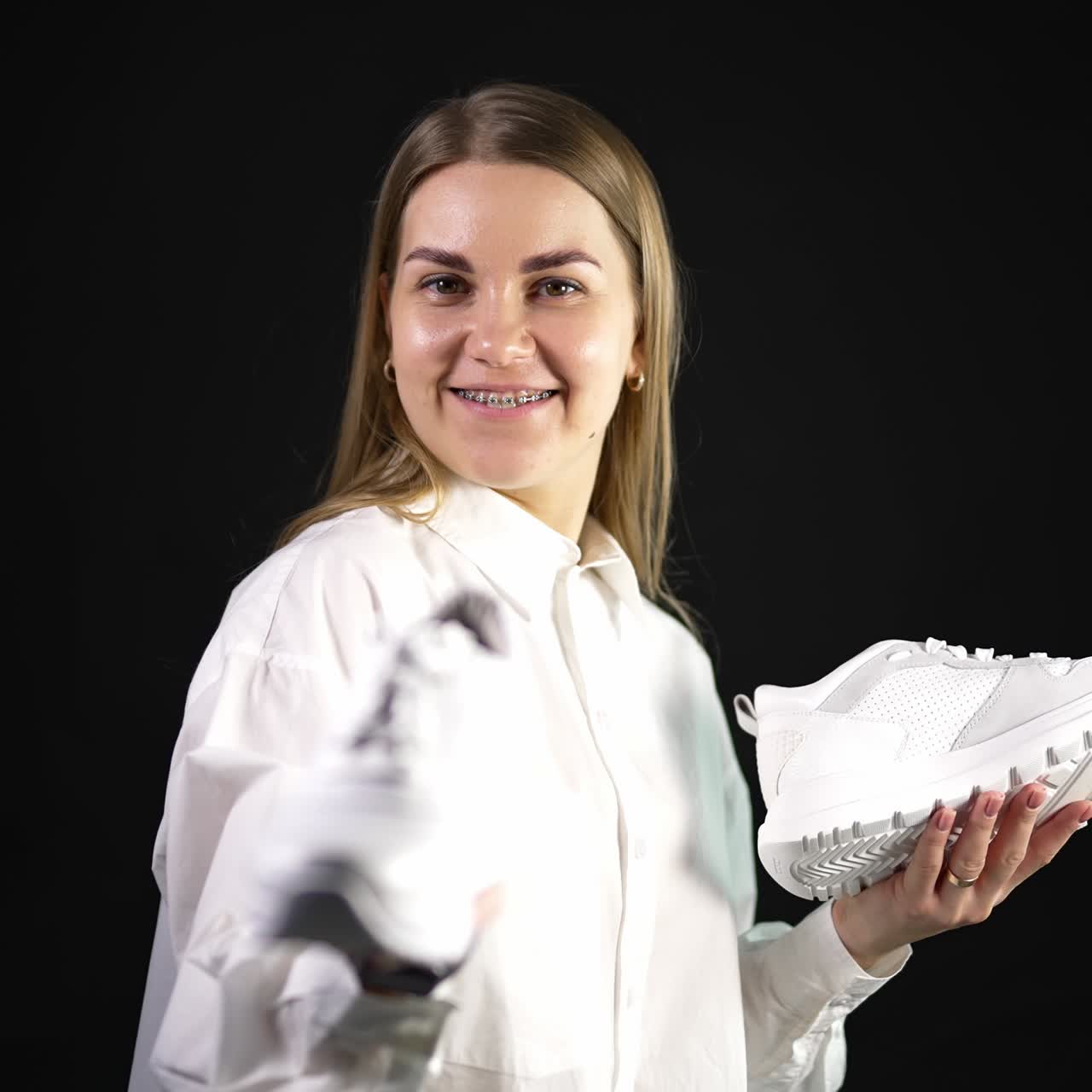 Lady in white shirt holds white sneakers from different pairs in both hands. Woman is making choice between similar shoes. Black backdrop