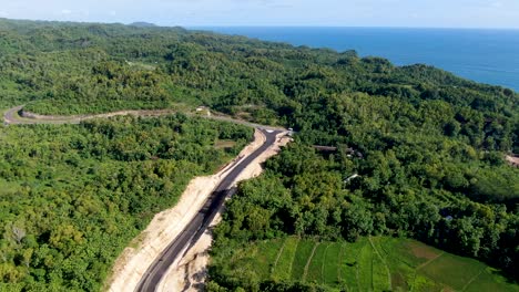 blue ocean water and winding road under construction in vibrant forest area of indonesia, aerial view