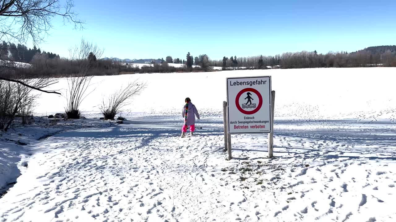 A child in a colorful scarf stands near a "Danger to Life" sign at the frozen Rottachsee, warning of thin ice and prohibiting access. Snowy landscape with bare trees and a clear blue sky. Rottachsee.