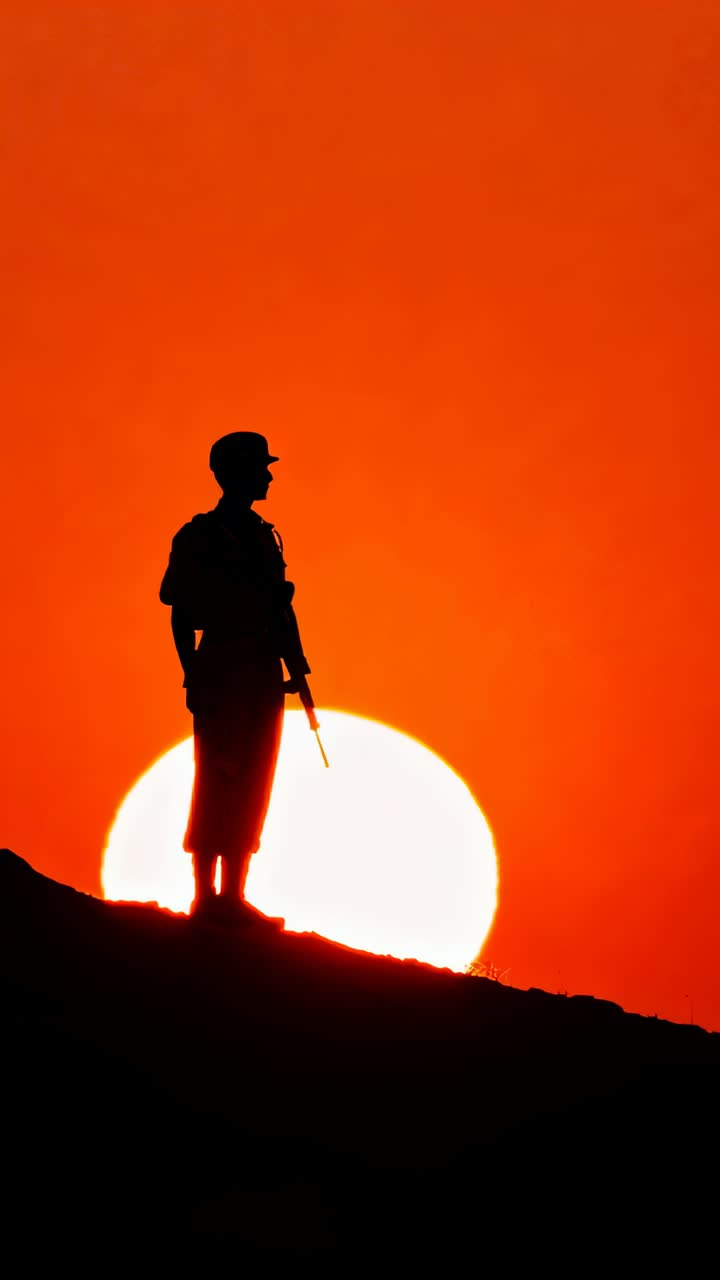 Video verticale: Soldato in piedi che fa la guardia su una cresta mentre il sole tramonta, con fucile, elmetto, zaino e stivali.