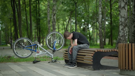 un joven con una camiseta negra se sienta con la cabeza hacia abajo, perdido en sus pensamientos, en un banco del parque, a su lado está su bicicleta al revés, y en la distancia borrosa, la gente está ocupada en el fondo