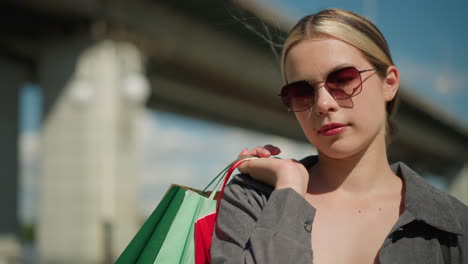 white woman wearing grey shirt and sunglasses holds several colorful shopping bags backwards outdoor , she stands confidently while the background is slightly blurred, featuring a bridge