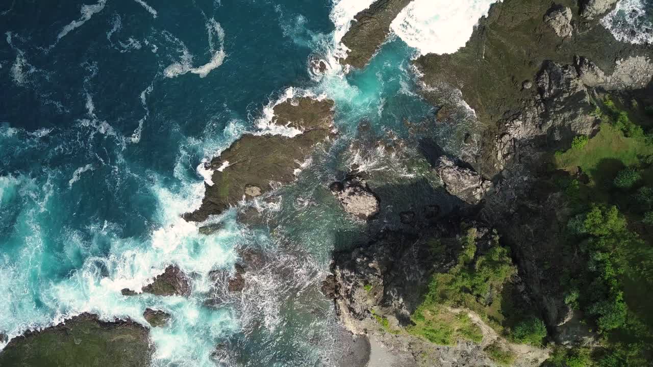 Ripresa aerea scorrevole delle onde dell'Oceano Indiano che colpiscono massi e barriera corallina sulla spiaggia in una giornata di sole, con una collina ricoperta di alberi visibile a lato - Collina di Pengilon, Indonesia, Asia
