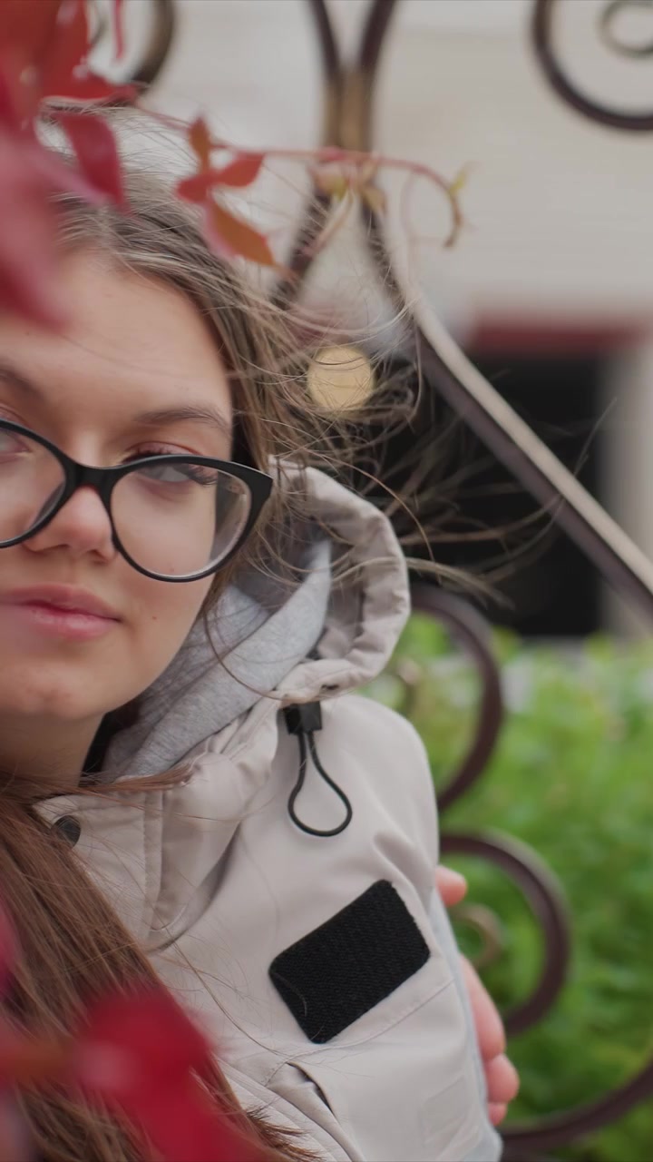 Profilo di un individuo con capelli castani che indossa occhiali. Un leggero fruscio di vegetazione dietro la ragazza suggerisce un ambiente esterno. La scena include verde misto a fogliame colorato.