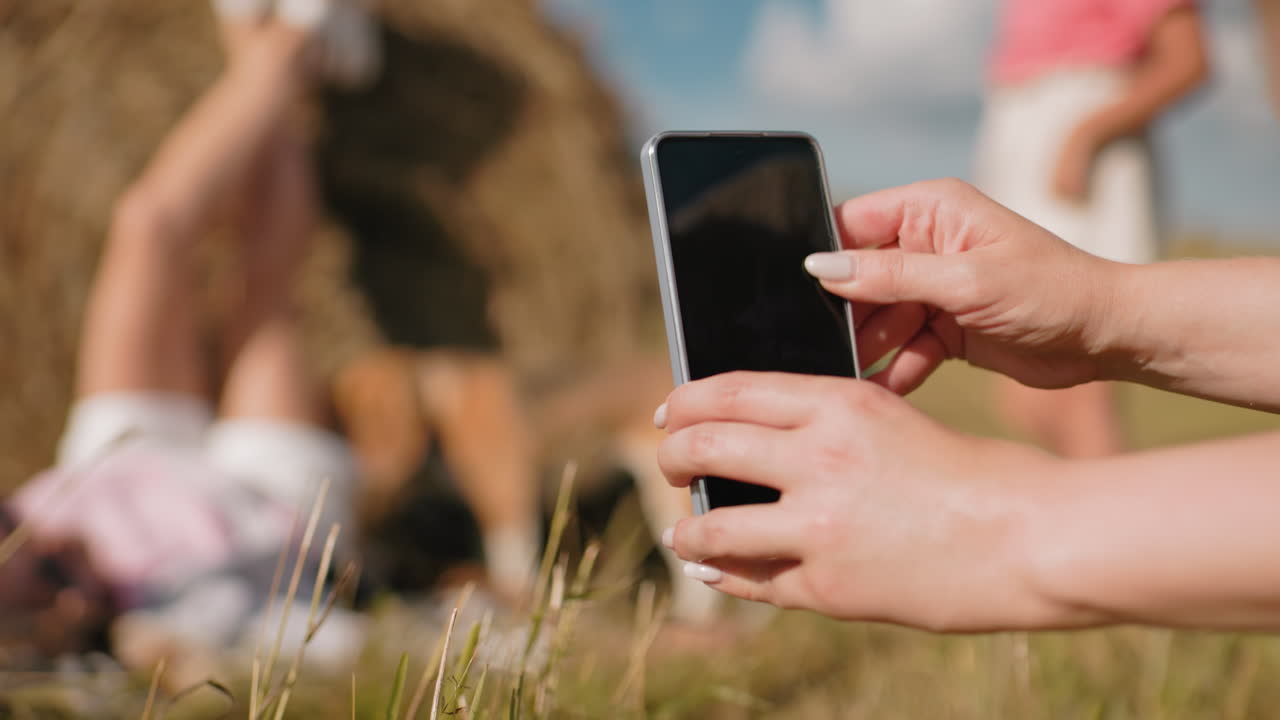 primer plano de una persona con un teléfono inteligente tomando una foto de un sujeto tendido en una bala de heno en un campo iluminado por el sol, con un fondo borroso de escena relajante, atmósfera de verano y tonos dorados cálidos