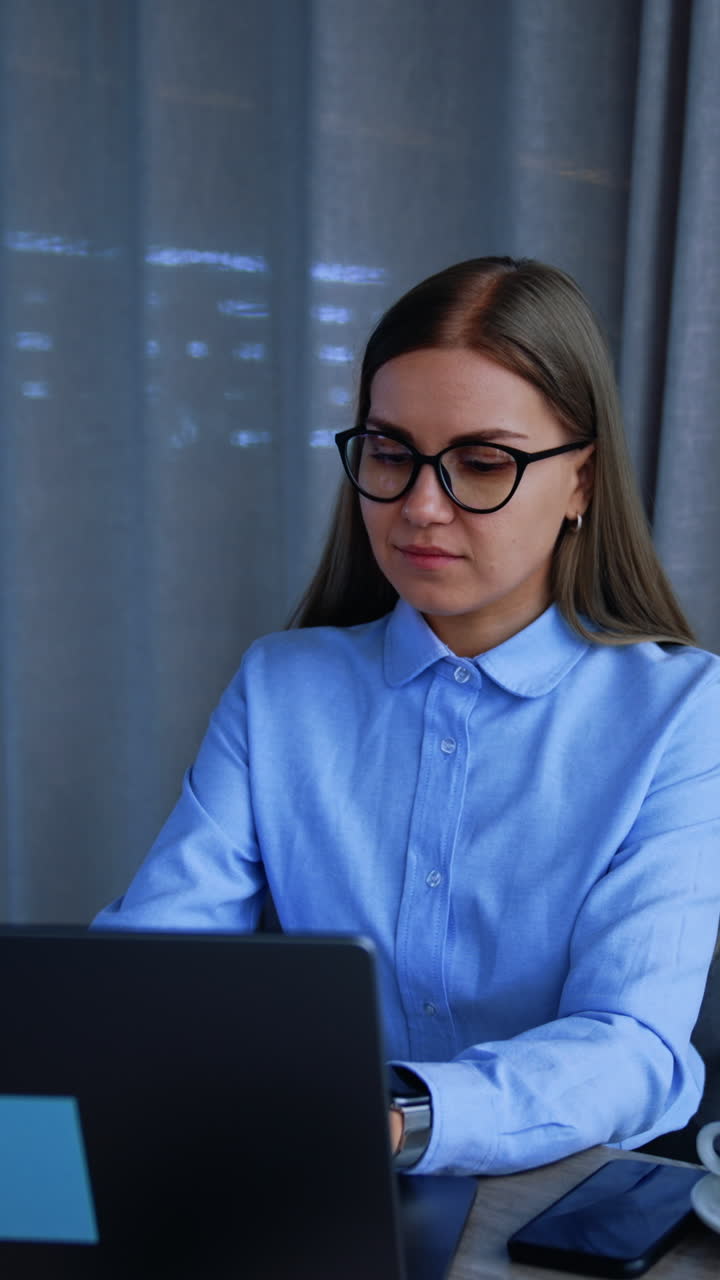 Beautiful young woman in glasses works on her laptop. Lady stands up, takes her cup and walks to the panoramic window in office. Vertical video