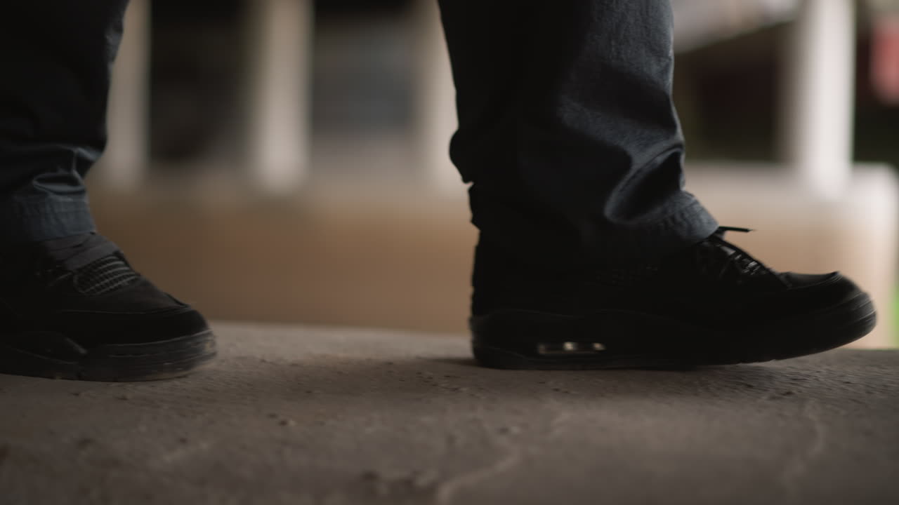 Scarpe da ginnastica nere su una sporgenza di cemento, primo piano dal basso che mostra suole rovinate e pantaloni di jeans, passi lenti e cauti vicino a un pilastro in un sottopassaggio buio, luci bokeh ambientali sullo sfondo, atmosfera urbana grintosa.
