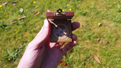 Close-up-of-a-hand-opening-squared-wooden-pocket-compass-in-a-park-with-grassy-background