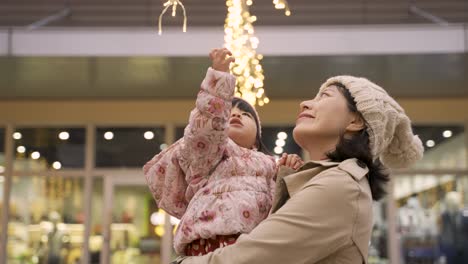 slow motion of cute curious asian toddler baby girl raising up hand trying to touch outdoor christmas lights in her mother’s arms outside an urban shopping mall