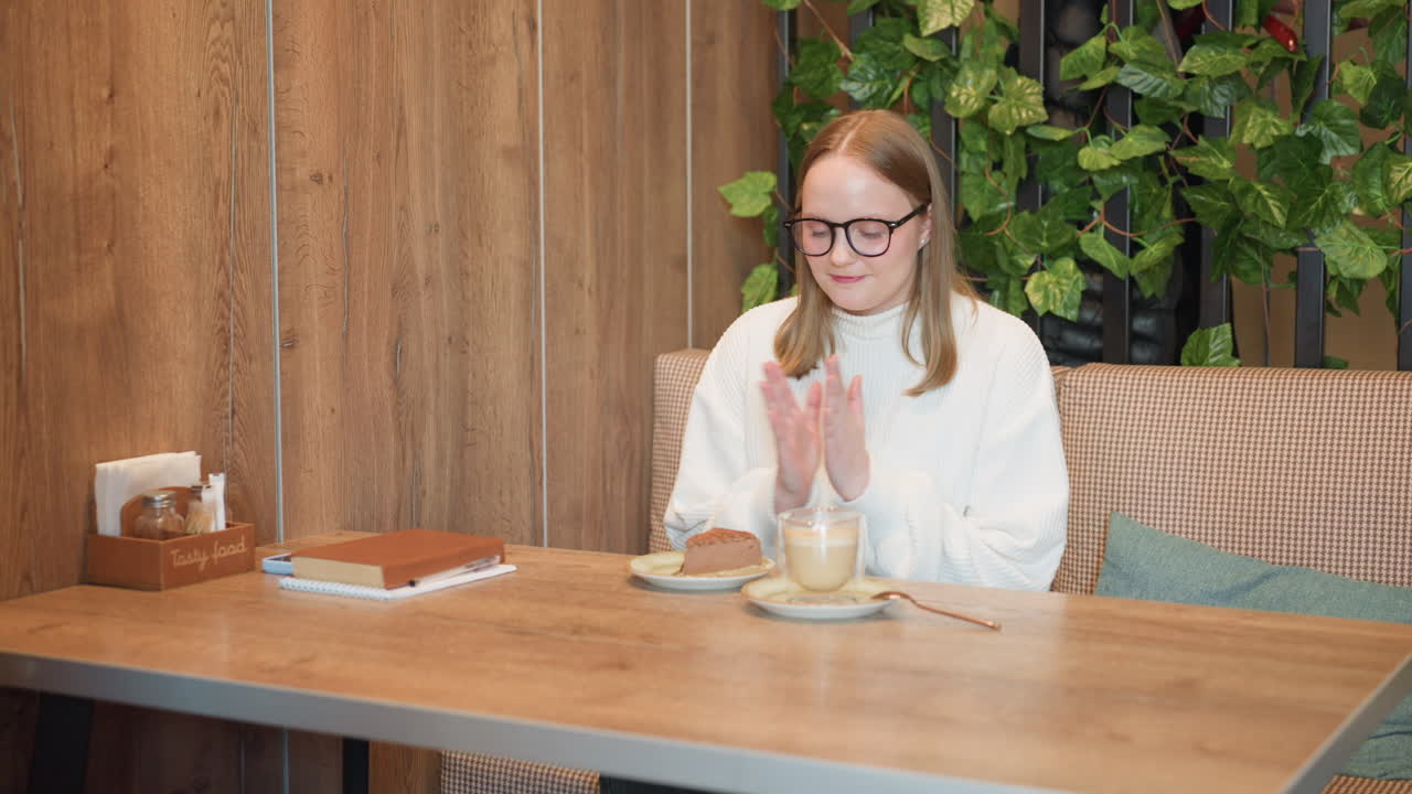 Joyful woman in white sweater smiles warmly and clasps hands in front of dessert and drink on wooden cafe table, expressing gratitude or excitement before eating in cozy, warmly lit indoor setting