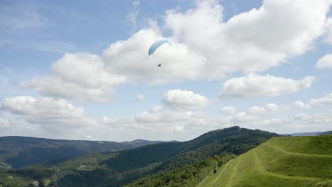 Tres-Parapentes-Que-Vuelan-Sobre-Colinas-Verdes,-Un-Parapente-Azul-Claro-Gira-En-El-Centro-Del-Marco-Contra-Un-Cielo-Azul-Con-Nubes-Giratorias