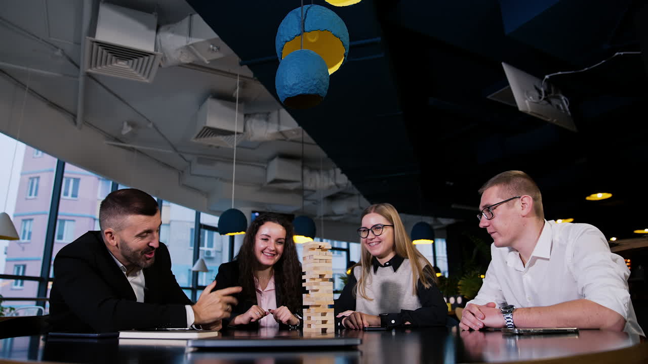 Four adults play table games sitting in the café. People playing jenga, laughing, having great time together.