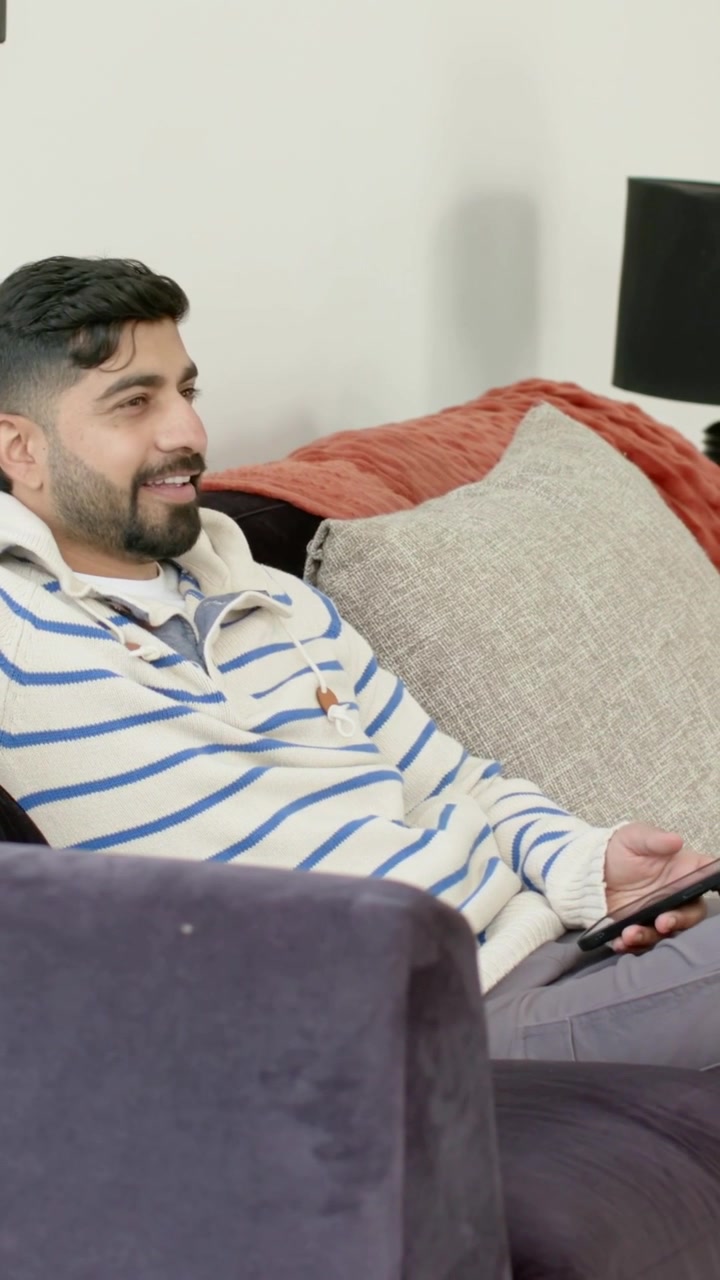 Video verticale: Un uomo con un maglione a righe tiene in mano uno smartphone, toccando e scorrendo lo schermo sul divano di casa.