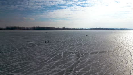 Fishing-and-walking-on-frozen-lake-in-Mitchell's-Bay,-St-Clair-Lake-with-windmills-in-the-background,-60fps-aerial-drone-shot