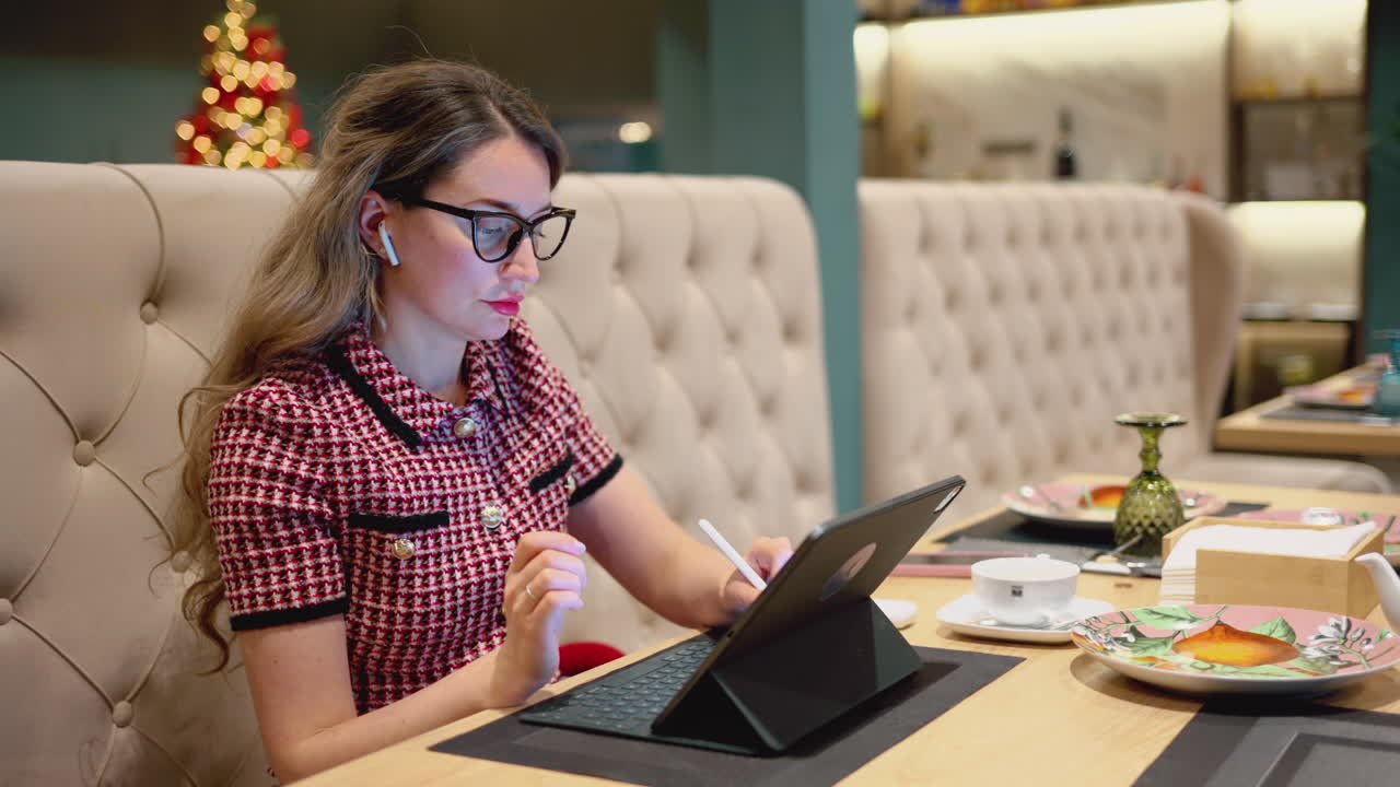 Brunette woman with glasses working on a tablet with wireless earphones at a restaurant, with a Christmas tree on the background