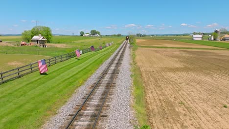 Una-Vista-Aérea-De-Un-Tren-De-Vapor-Que-Se-Acerca-Lentamente-A-Través-De-Tierras-De-Cultivo-Y-Una-Valla-Con-Muchas-Banderas-Americanas-Ondeando-En-El-Viento-En-Un-Soleado-Día-De-Primavera