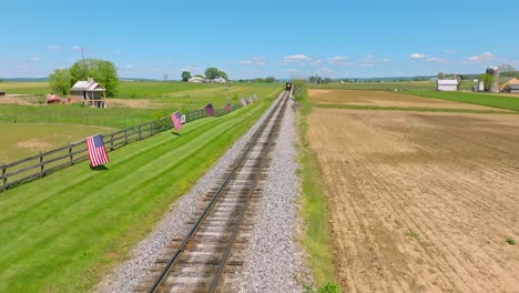 an aerial view of a steam train approaching slowly thru farmlands and a fence with many american flags waving in the wind on a sunny spring day