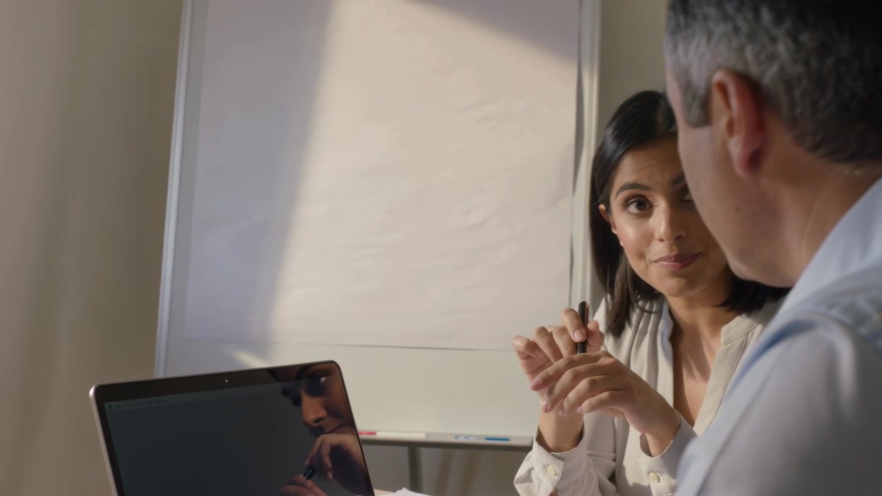Lifting pen, woman in light blouse, man in shirt reviewing report at office, laptop between them