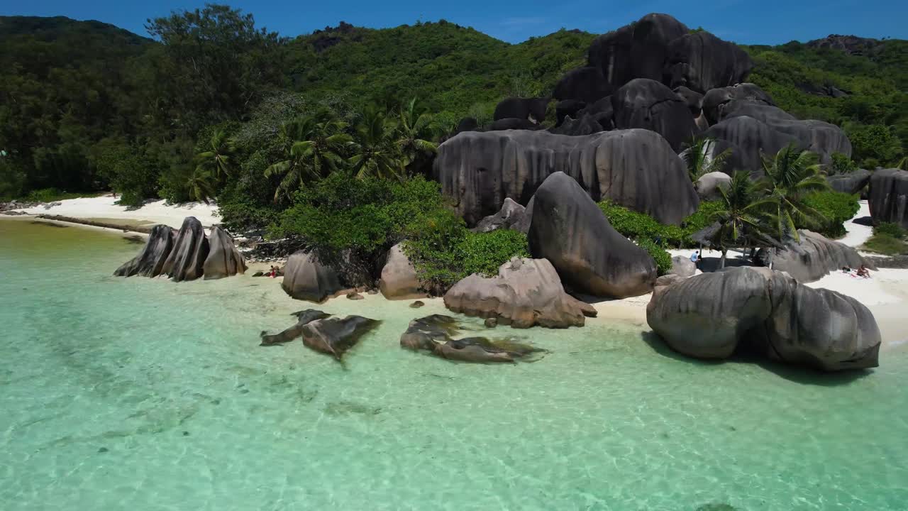 Pemandangan udara melayang Pantai Anse Source D'argent di La Digue, Seychelles, dengan batu-batu besar, pohon palem, dan air tropis biru jernih.