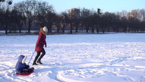 family,-sledding,-season-and-people-concept---happy-mother-pulling-on-ice-sled-with-child-outdoors-in-winter