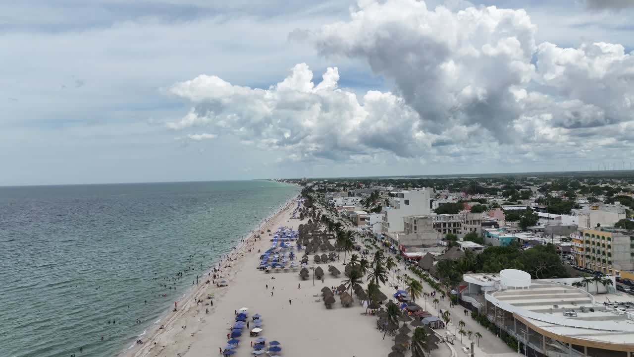 Drone flying forward along the crowded beach of Progreso, Yucatán, showing turquoise water, umbrellas, and the city under dramatic clouds