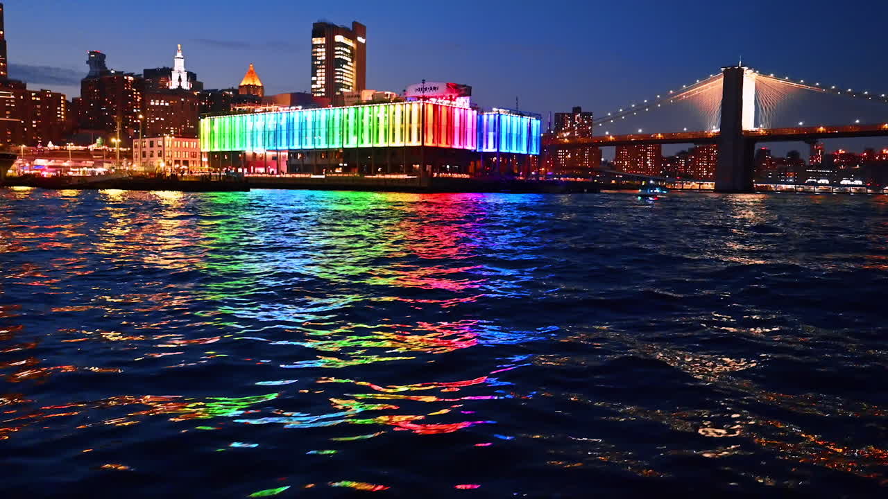 Beautiful rainbow illumination of Pier 17 Bar reflecting in the dark riverscape. River cruise approaching the Brooklyn Bridge at night. New York, USA