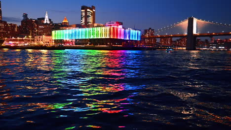 beautiful rainbow illumination of pier 17 bar reflecting in the dark riverscape. river cruise approaching the brooklyn bridge at night. new york, usa